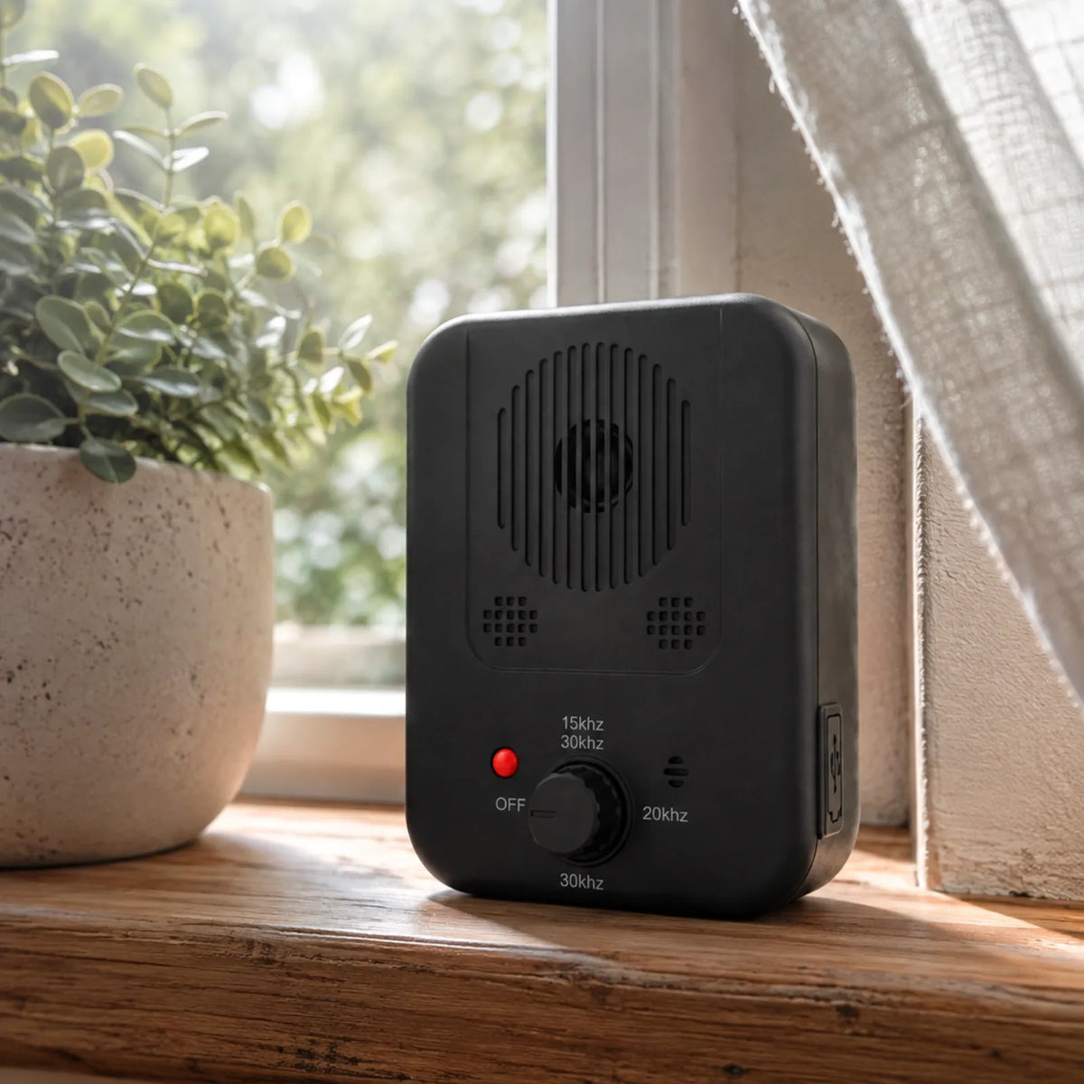 A black electronic signal jammer device with a rotary frequency control knob and red power indicator light, sitting on a wooden windowsill next to a potted plant.