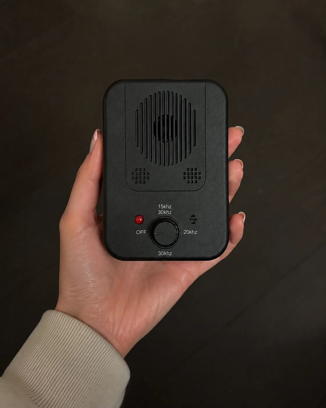 A hand holding a small black electronic device with a speaker grille, frequency controls, and a red indicator light against a dark wooden background.